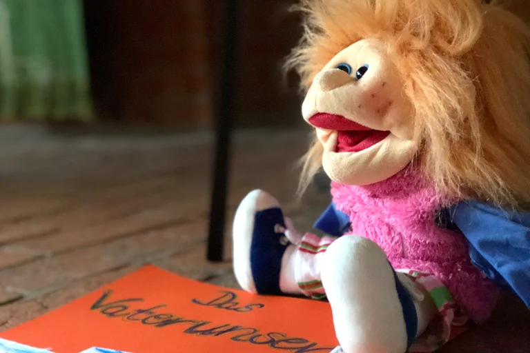 Eine Handpuppe mit langen Haaren, sitzt rechts in der Ecke auf einem roten Pappschild, auf welchem "Das Vaterunser" steht